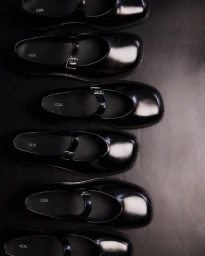 Overhead arrangement of six black patent Mary Jane shoes with chunky lug soles lined up against a dark backdrop for COS.
