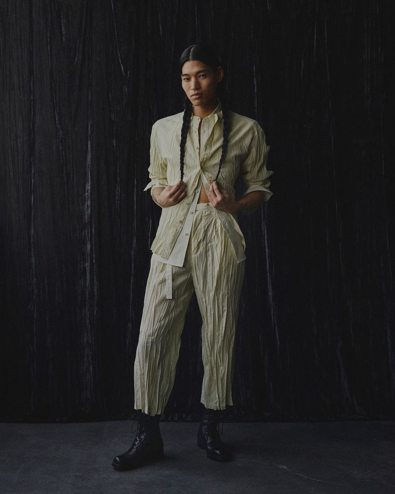 Non-binary model in wrinkled light yellow collared shirt, pants, and black boots against dark textured backdrop in a fashion shoot.