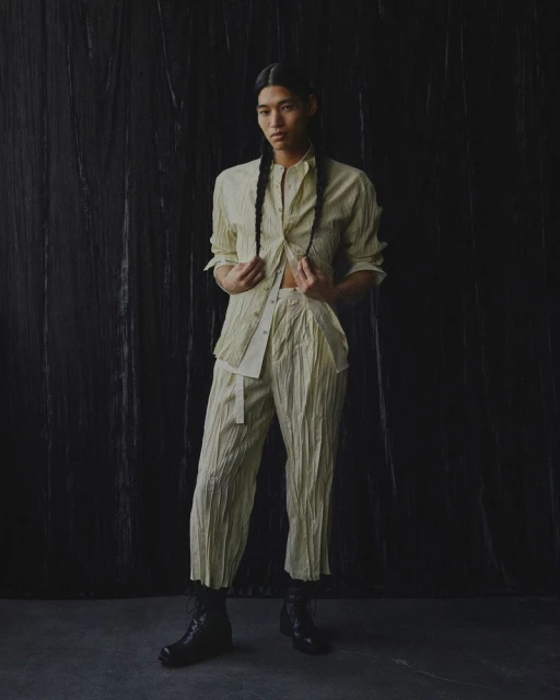 Non-binary model in wrinkled light yellow collared shirt, pants, and black boots against dark textured backdrop in a fashion shoot.