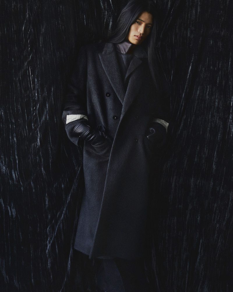 Fashion shot of a person with long dark hair wearing a gray overcoat, dark top, and gloves against a textured dark background.