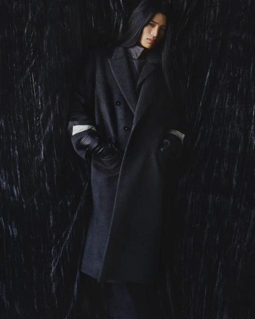 Fashion shot of a person with long dark hair wearing a gray overcoat, dark top, and gloves against a textured dark background.