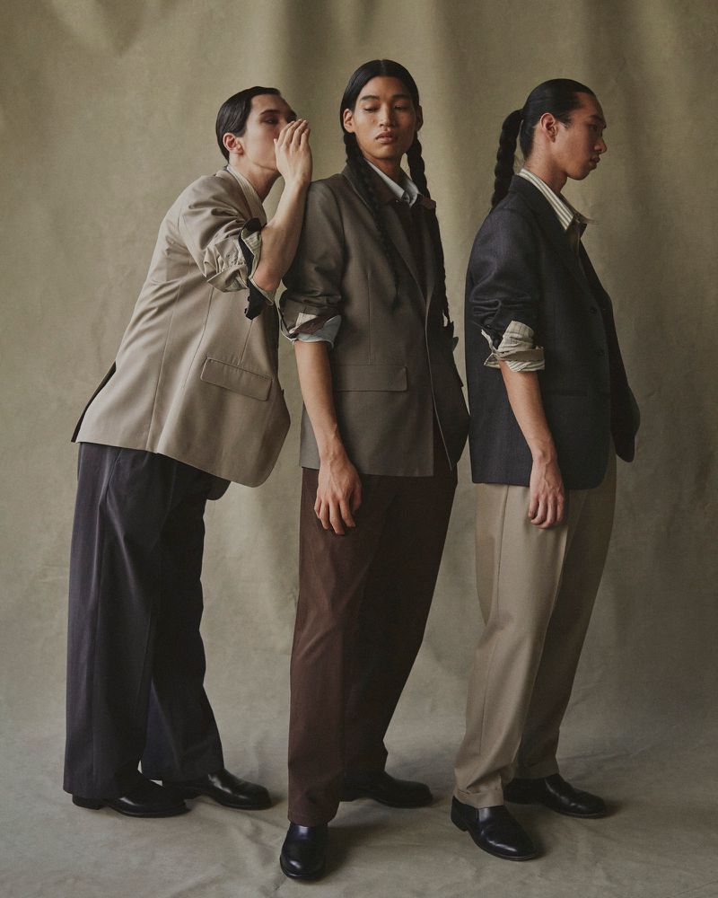 Three people in formal attire pose indoors with one person whispering to another, while the third person stands looking away.