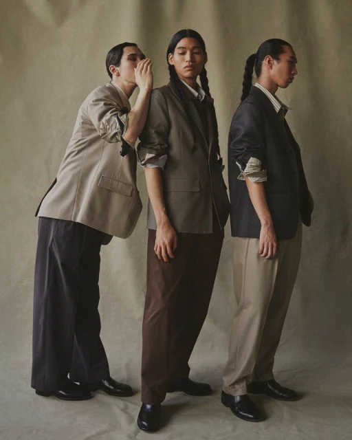 Three people in formal attire pose indoors with one person whispering to another, while the third person stands looking away.