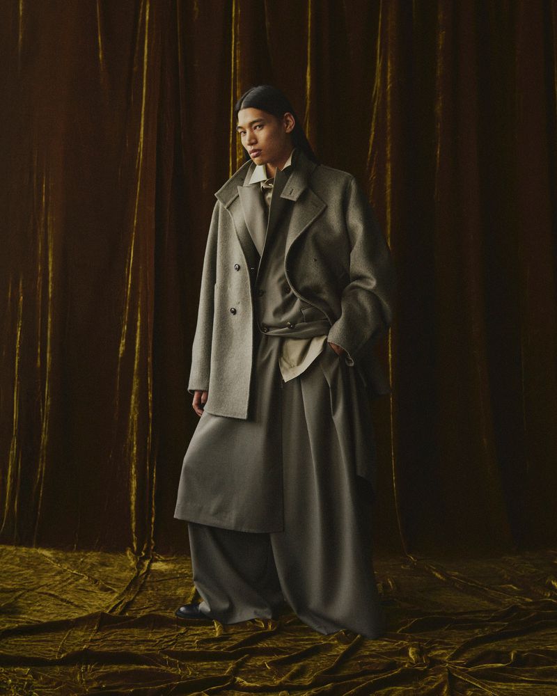 Woman poses indoors wearing a light-gray double-breasted wool coat, trousers, and blouse against an olive-green velvet backdrop.