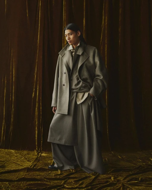 Woman poses indoors wearing a light-gray double-breasted wool coat, trousers, and blouse against an olive-green velvet backdrop.
