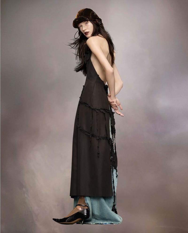 A woman with dark hair wearing a dark dress and hat poses in a studio with a neutral background. The woman looks toward the camera.