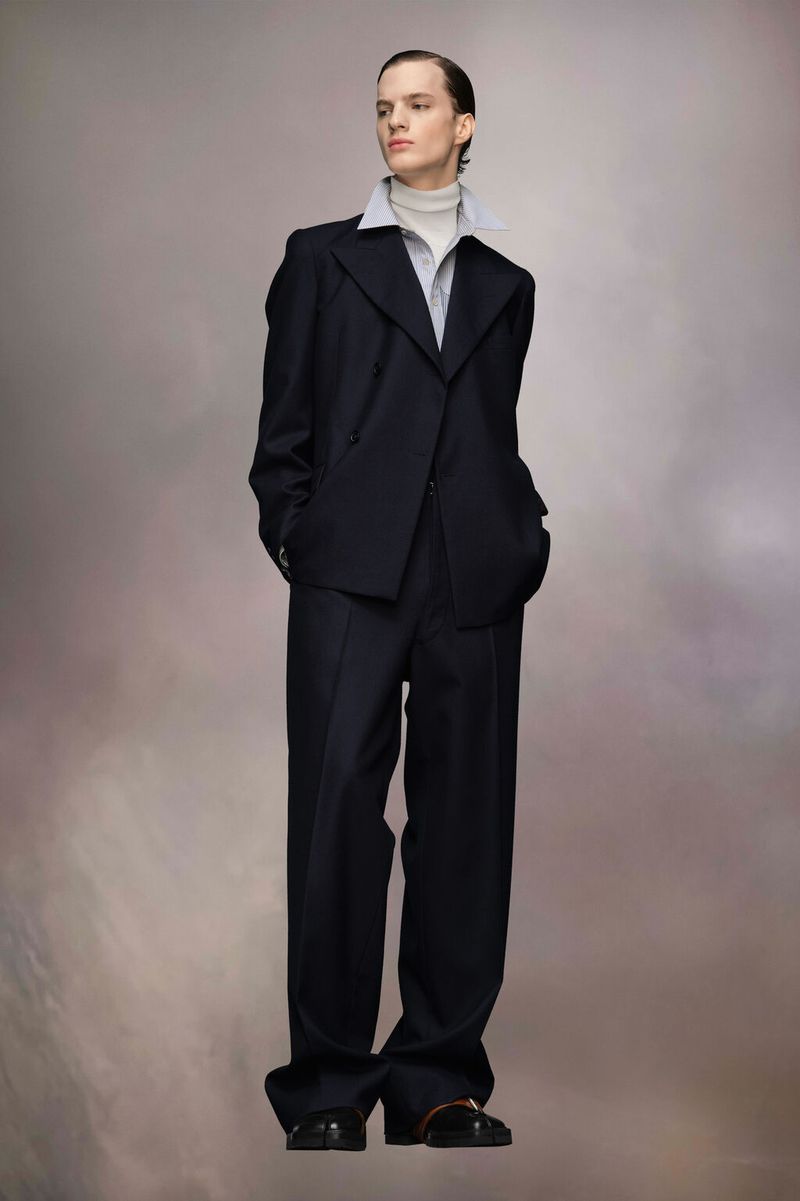 Model wearing a dark oversized blazer and trousers with a layered collar shirt against a neutral gradient background