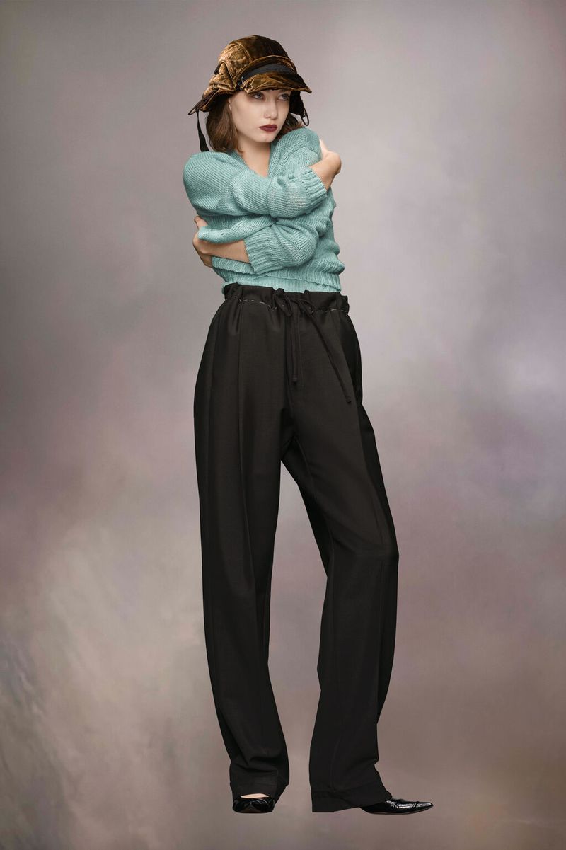 Full-shot of a woman in a light-blue sweater, dark pants, and a brown hat. She's embracing herself against a muted-brown backdrop.