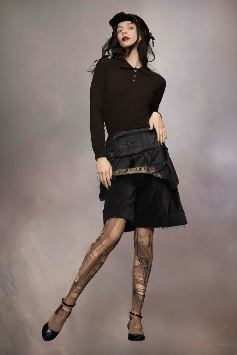 A woman stands indoors wearing a dark long-sleeved polo shirt, ornate shorts, patterned tights, and black patent leather Mary Jane heels.