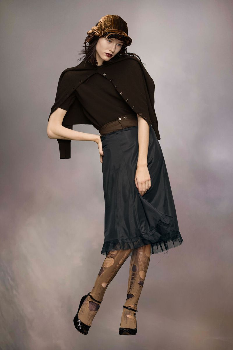 A fashionably dressed woman poses in a brown hat, cardigan, t-shirt, and semi-sheer black skirt against a neutral backdrop in a studio shot.