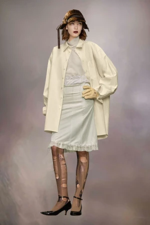 Full-length fashion shot of a woman wearing a tan bucket hat, oversized beige shirt jacket, and a pale skirt with distressed tights.
