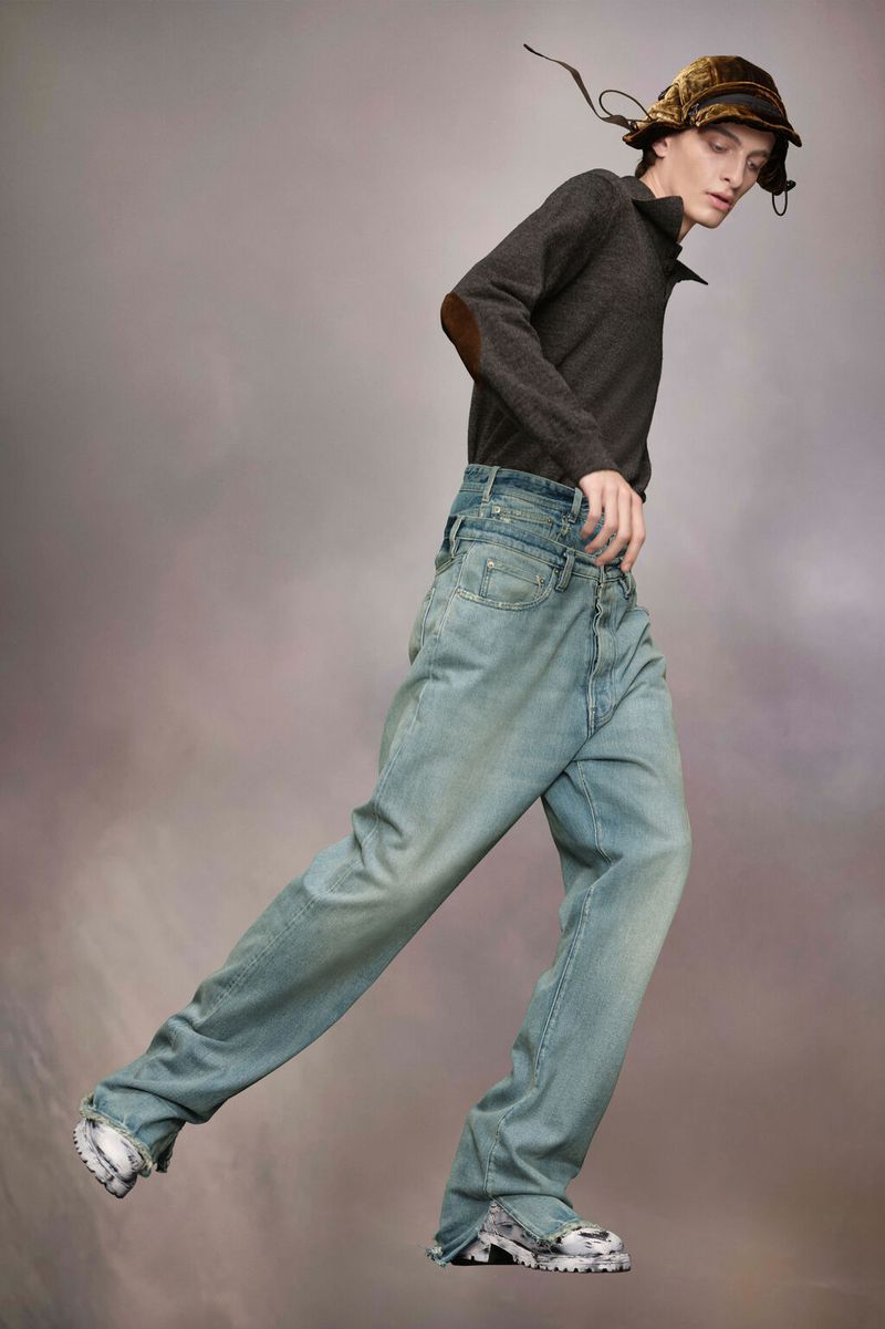 Fashion shot of a slender male model in light-wash, wide-legged denim jeans, dark top with elbow patches, and patterned hat in studio.