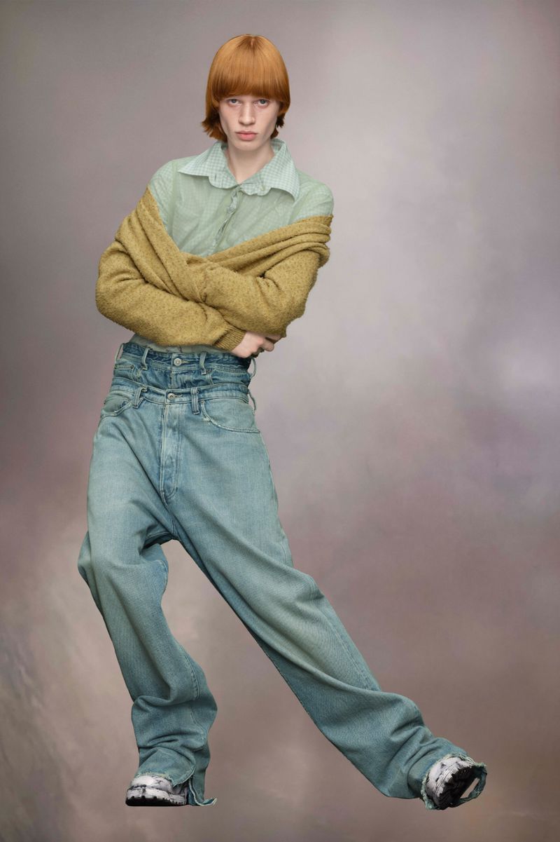 Person poses in a light blue shirt and oversized jeans with a gold sweater draped over their shoulders against a studio backdrop.