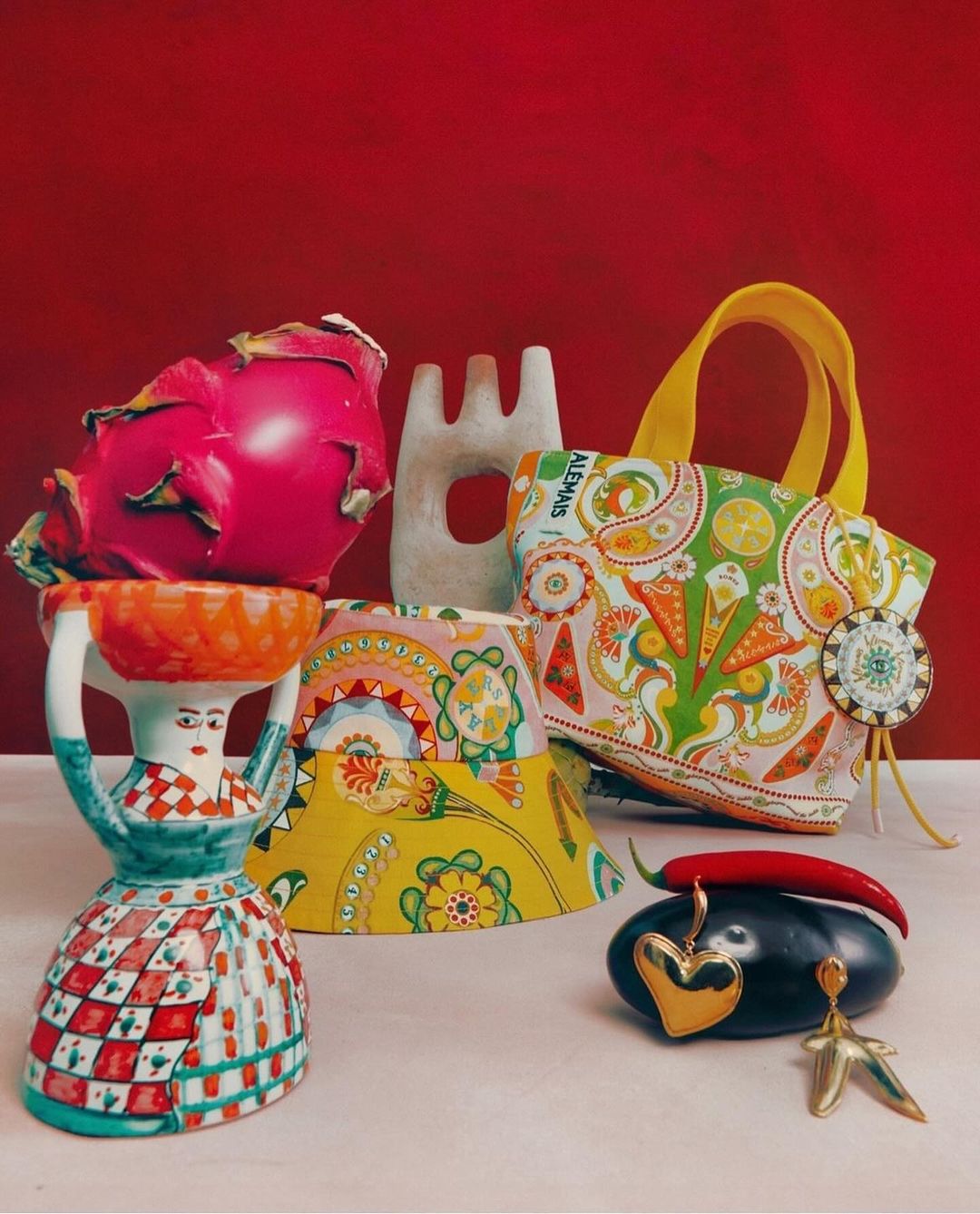 Still life photo of a dragon fruit on top of a woman-shaped vase, an abstract sculpture, a colorful tote bag, and jewelry.