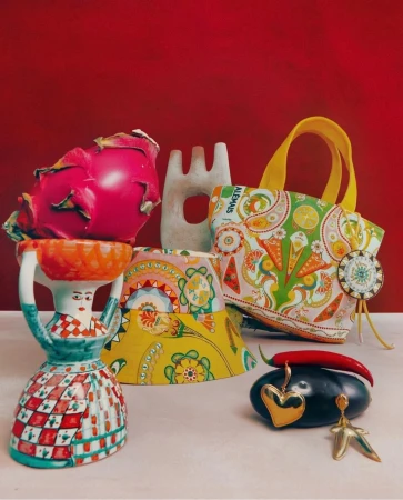 Still life photo of a dragon fruit on top of a woman-shaped vase, an abstract sculpture, a colorful tote bag, and jewelry.
