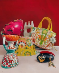 Still life accessories shoot featuring a patterned handbag, ceramic figurine, and dragon fruit against a red background.
