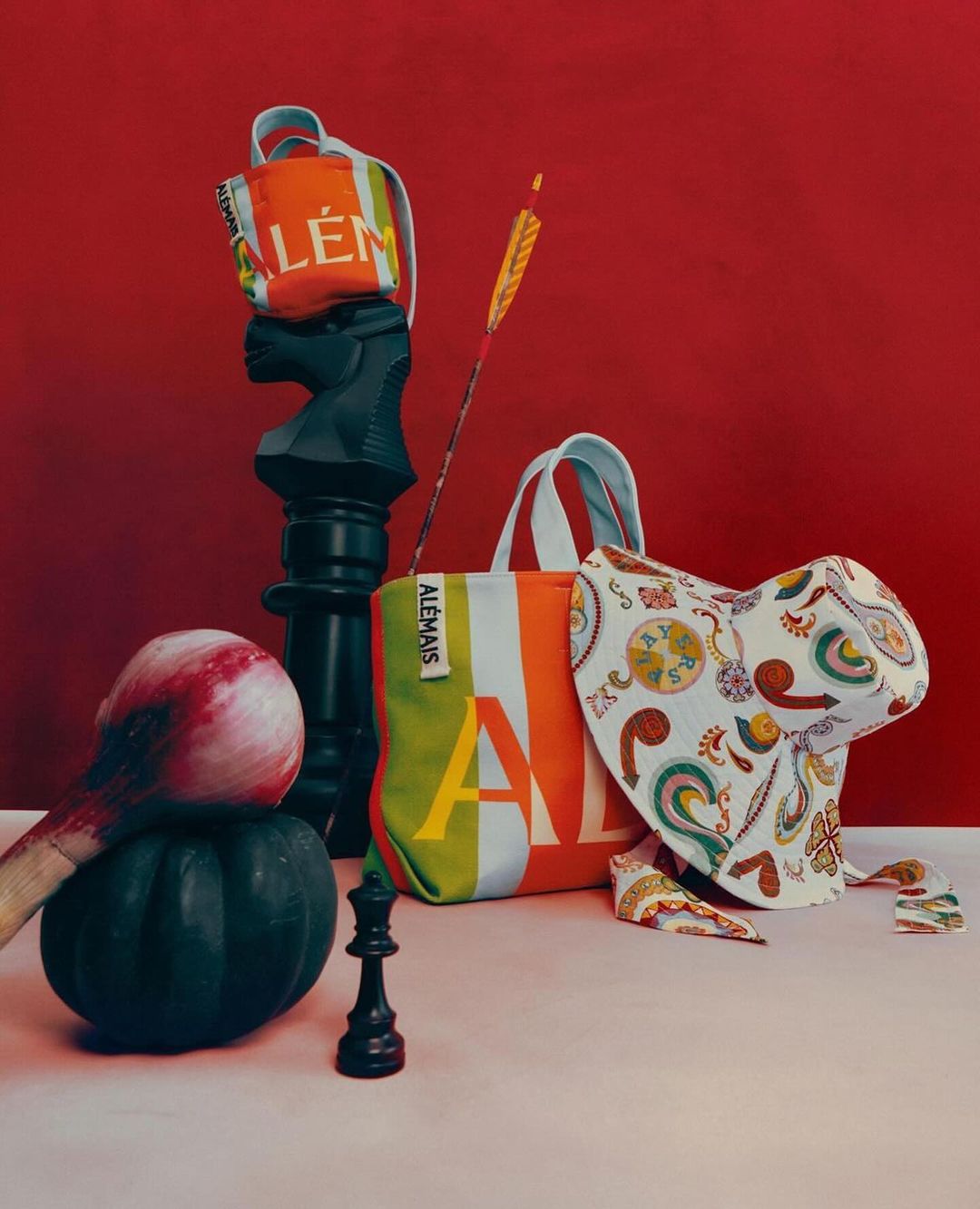 Still life composition featuring Alemais branded totes, chess pieces, an arrow, and unique objects against a solid red backdrop.