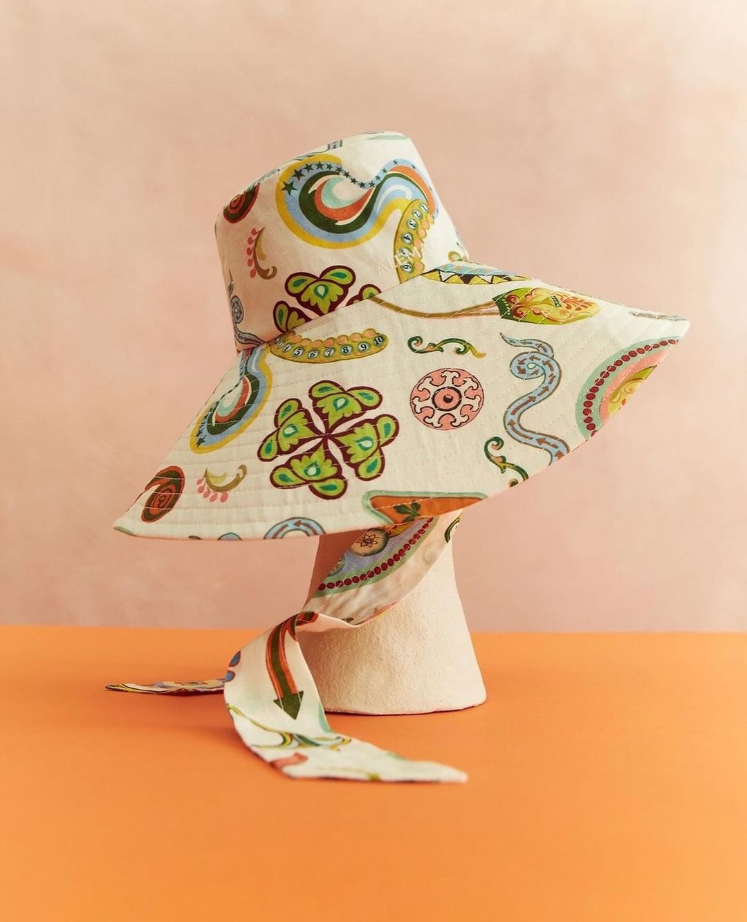Sun hat on mannequin head. A paisley-patterned bucket hat with a tie drapes on an off-white mannequin head against a peach and orange backdrop.