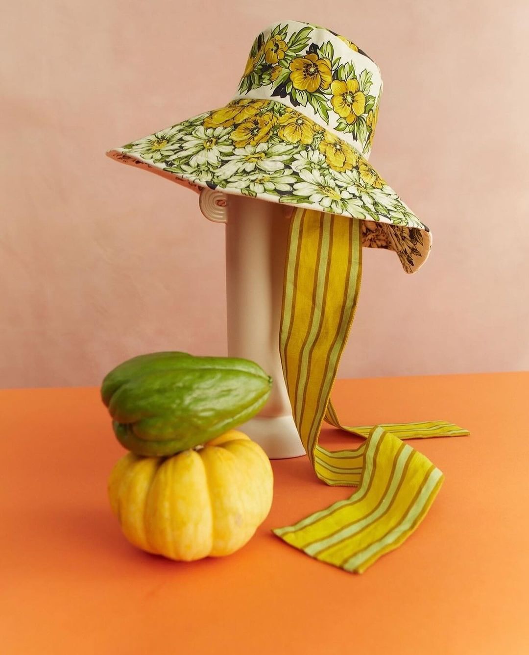 Floral bucket hat with a striped ribbon on a white vase. There are two gourds, one green and one yellow, on an orange surface.