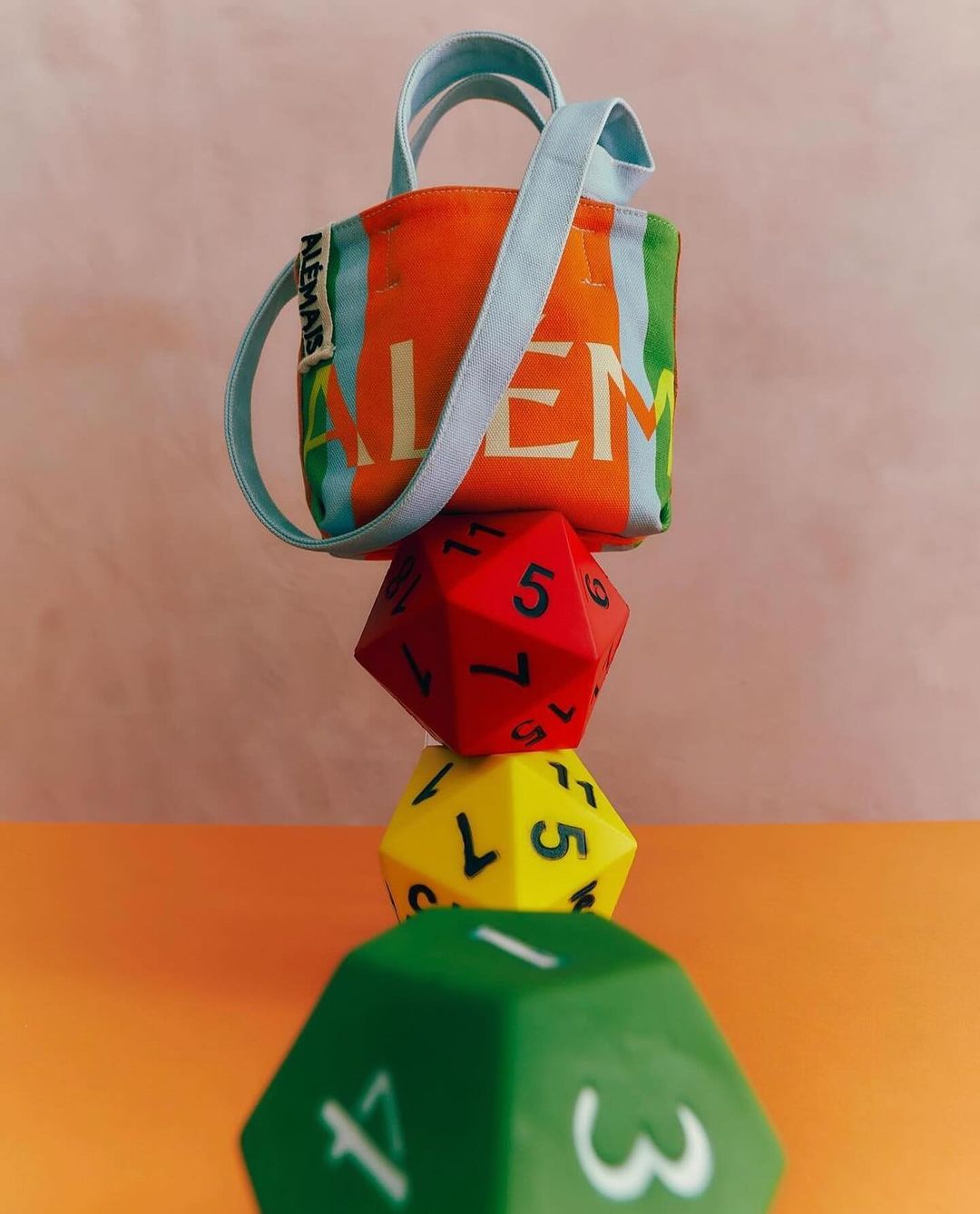Colorful Alemas brand tote bag stacked atop three colorful polyhedral dice in an eye-level, indoor product shot.