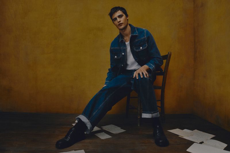 Adult male fashion model poses on a wooden chair in a blue denim jacket, dark wash jeans, and black boots, set against a yellow wall.