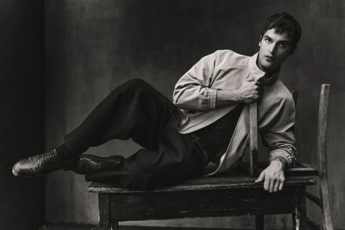Parker Van Noord reclining on a wooden table with a chair in a monochrome fashion portrait for Zara.