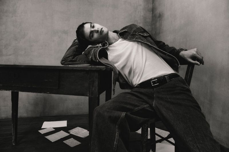 Monochrome fashion shot of a young adult man in a denim jacket and jeans, reclining on a chair at a desk with papers scattered on the floor.