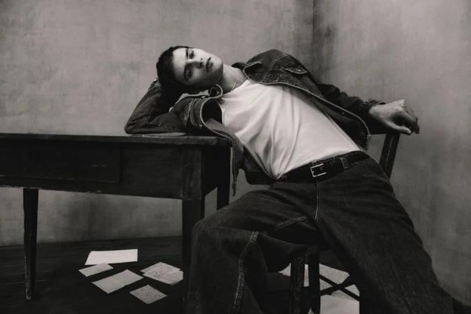 Monochrome fashion shot of a young adult man in a denim jacket and jeans, reclining on a chair at a desk with papers scattered on the floor.