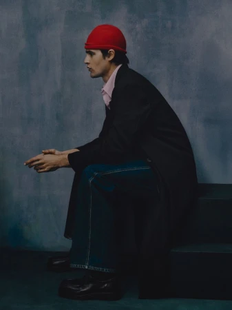 Model Parker Van Noord seated in profile wearing a red beanie, black coat, and denim jeans against a textured blue backdrop.