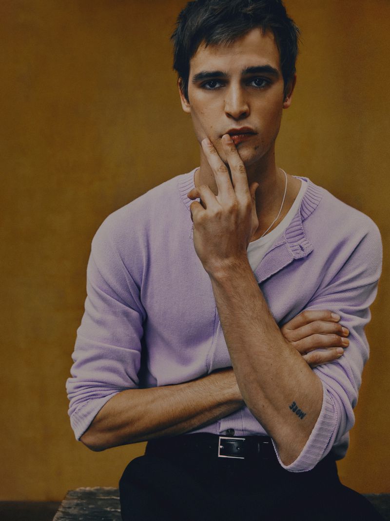 Portrait of a young adult man with dark hair wearing a lavender sweater and black pants, posing indoors against an orange background.