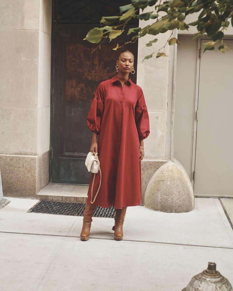 Elegant adult female in a red dress, accessorized with earrings, a purse, and boots, stands on a city sidewalk in front of a doorway.