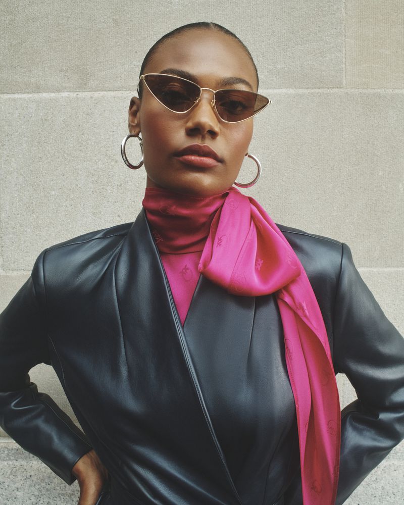 Fashionable African American woman poses outdoors in a black leather jacket, pink top, pink scarf, sunglasses, and hoop earrings.