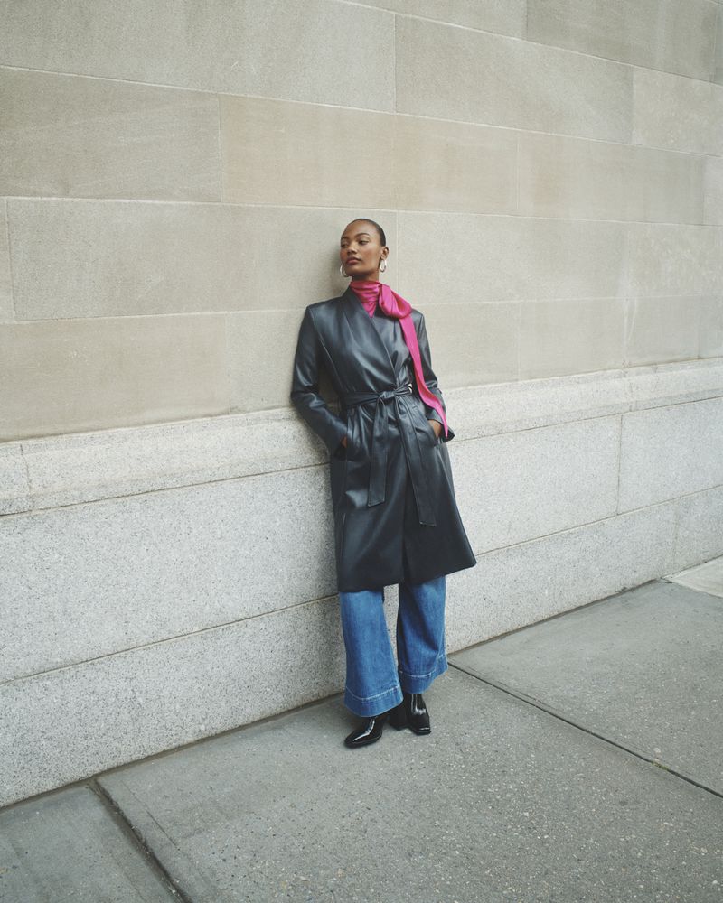 Model in dark leather coat and pink scarf leaning against a stone building wall