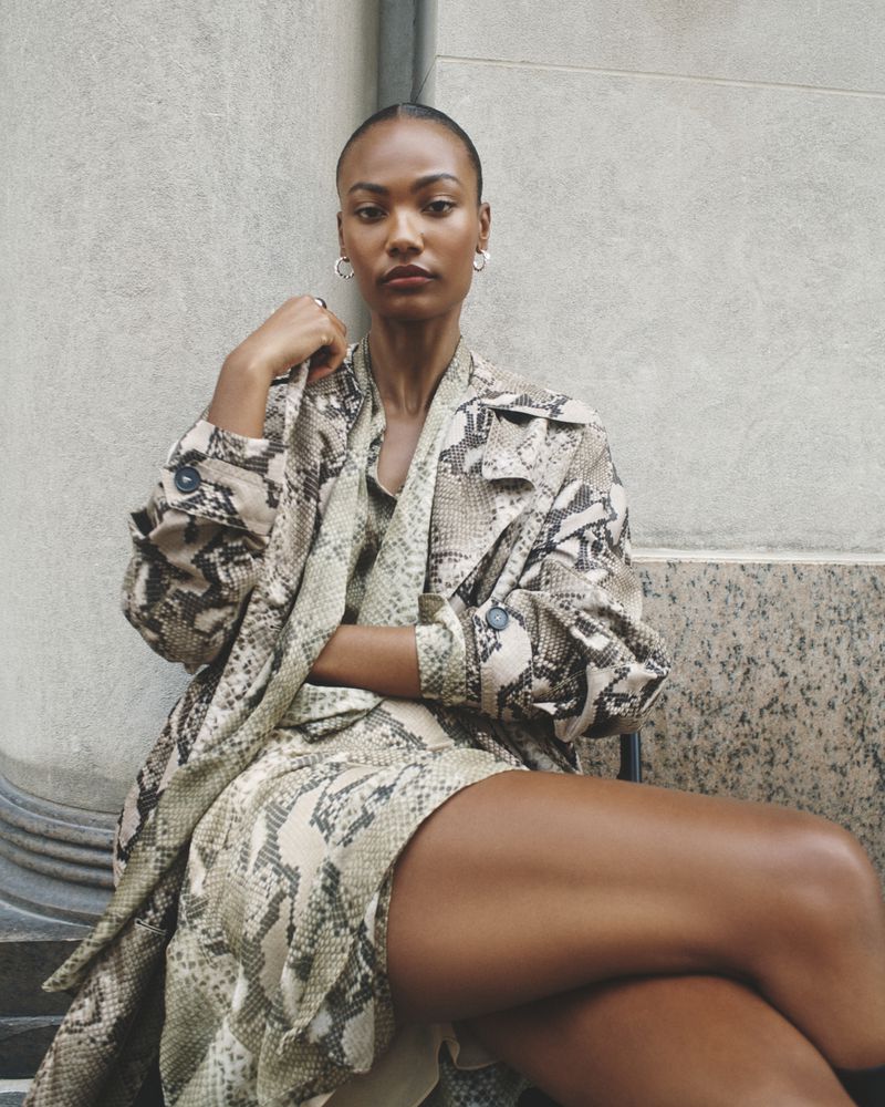Woman in snakeskin dress and coat sitting against gray wall. She is posing with arms crossed, looking at the camera.