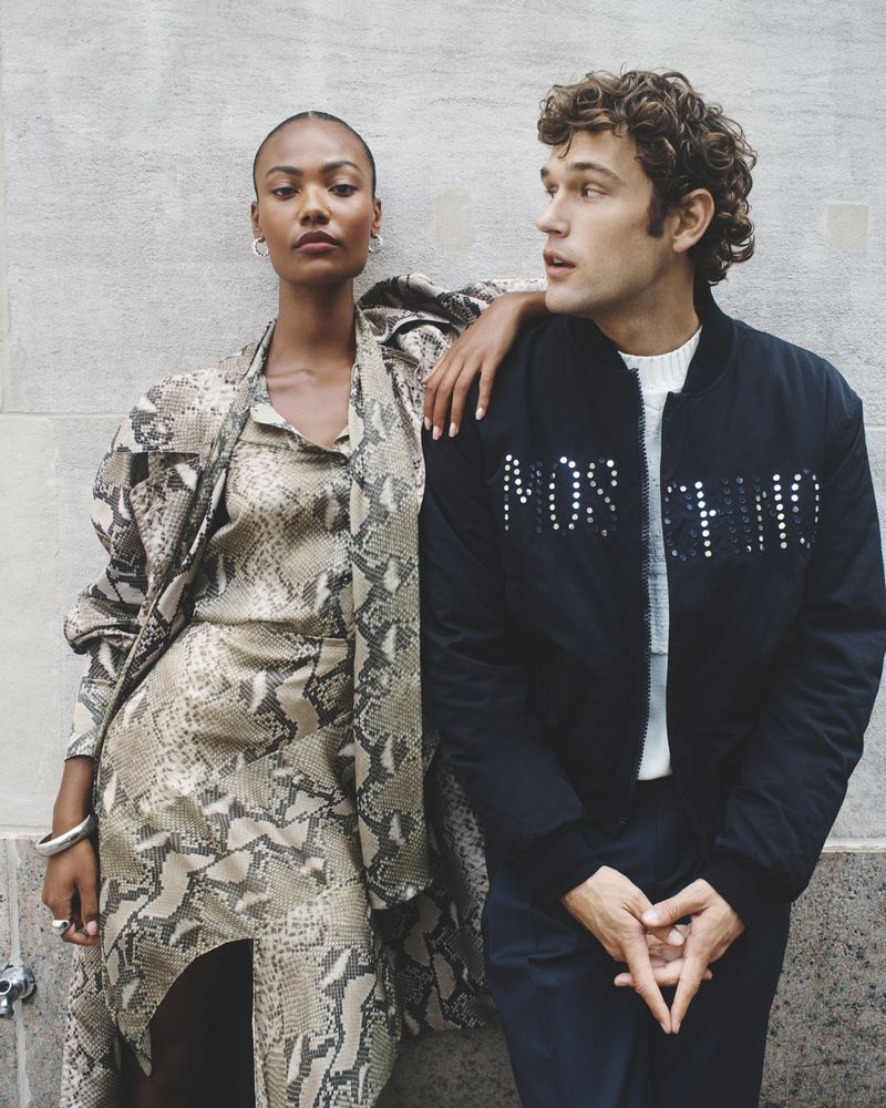 A couple poses against a neutral wall. The woman wears a snake-print dress with a jacket. The man wears a black bomber jacket with Moschino lettering.