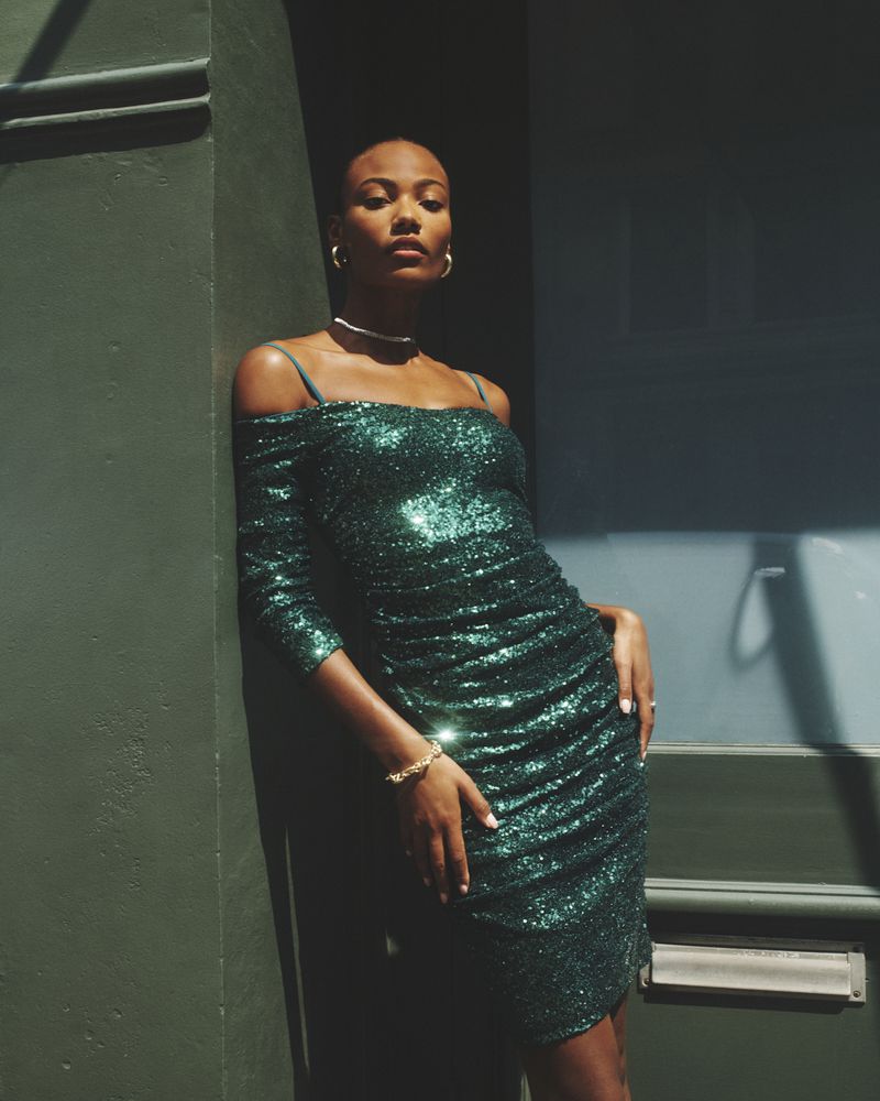 Attractive woman poses in a shimmering emerald green, off-the-shoulder mini dress against a green door, accented with gold bracelet and necklace.