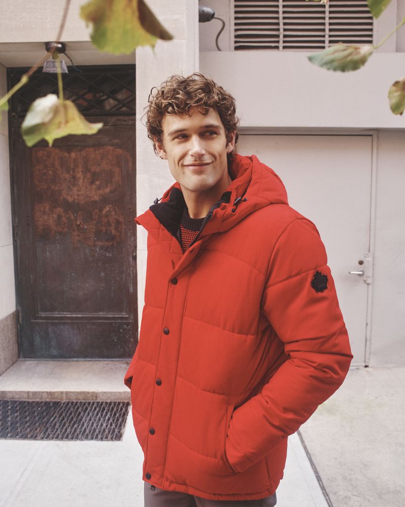 Man wearing a vibrant red puffer jacket standing outdoors near a building entrance with foliage framing the top foreground