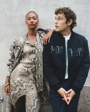 Two models leaning against a textured stone wall, wearing a snakeskin print outfit and a dark bomber jacket.