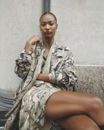 Model seated wearing a snakeskin print trench coat and matching dress against a neutral stone architectural background