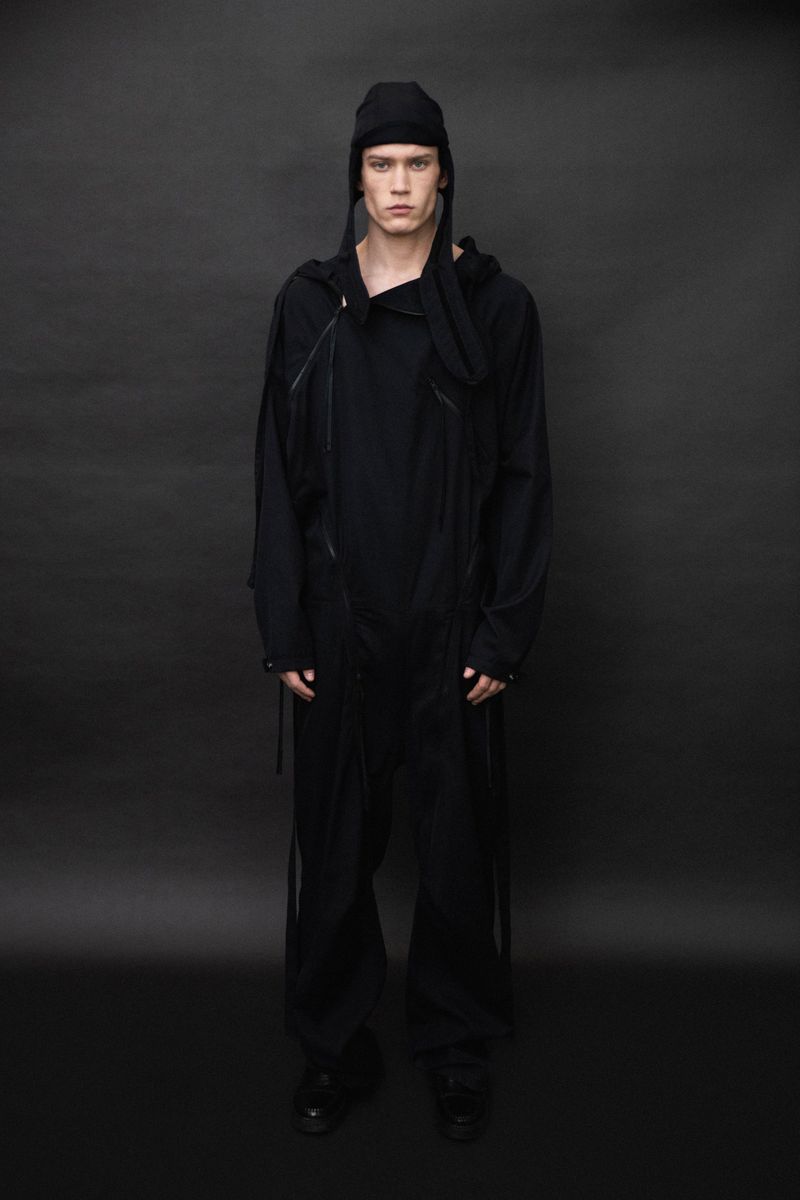 Fashionable adult male model poses in a black jumpsuit and beanie against a dark gray studio backdrop, showcasing a minimalist aesthetic.