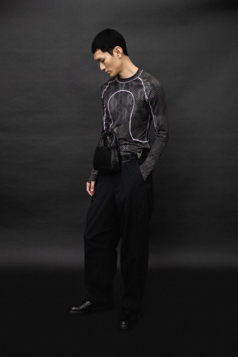 An Asian man in a black long-sleeved shirt and baggy pants poses in front of a dark backdrop, displaying fashion apparel and accessories.