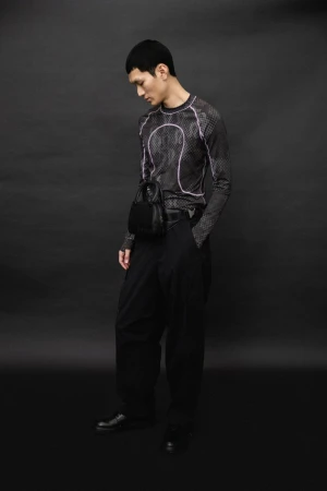 An Asian man in a black long-sleeved shirt and baggy pants poses in front of a dark backdrop, displaying fashion apparel and accessories.