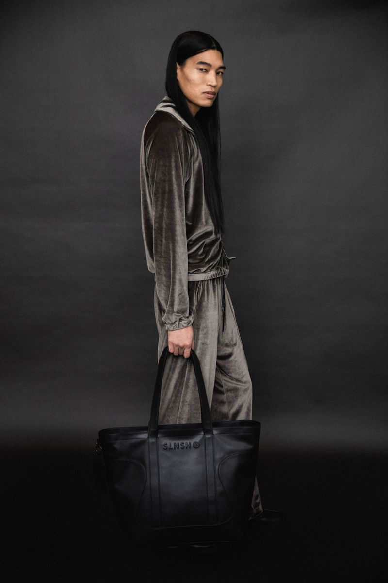An Asian male model poses in a grey velvet tracksuit, holding a black leather tote bag against a dark grey backdrop in a studio shot.