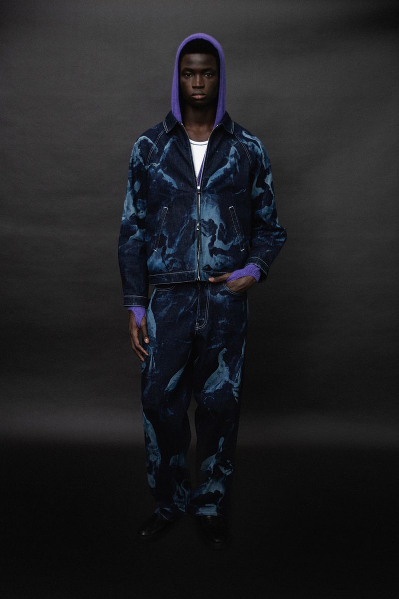 Dark-skinned adult man in matching patterned denim jacket and pants, wearing a purple hoodie, standing indoors against a grey background.