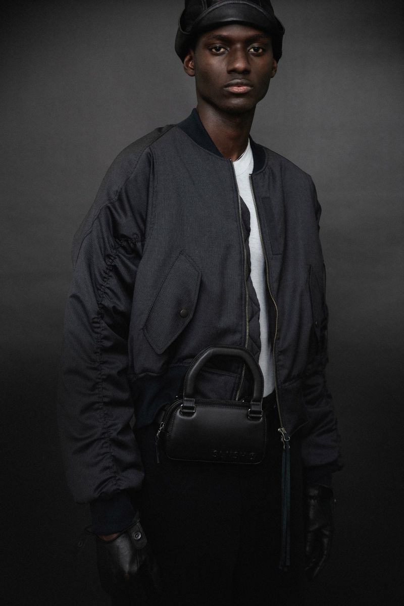 Fashionable adult man wearing a bomber jacket and a cap with a small handbag, posing in a studio with a dark grey background.