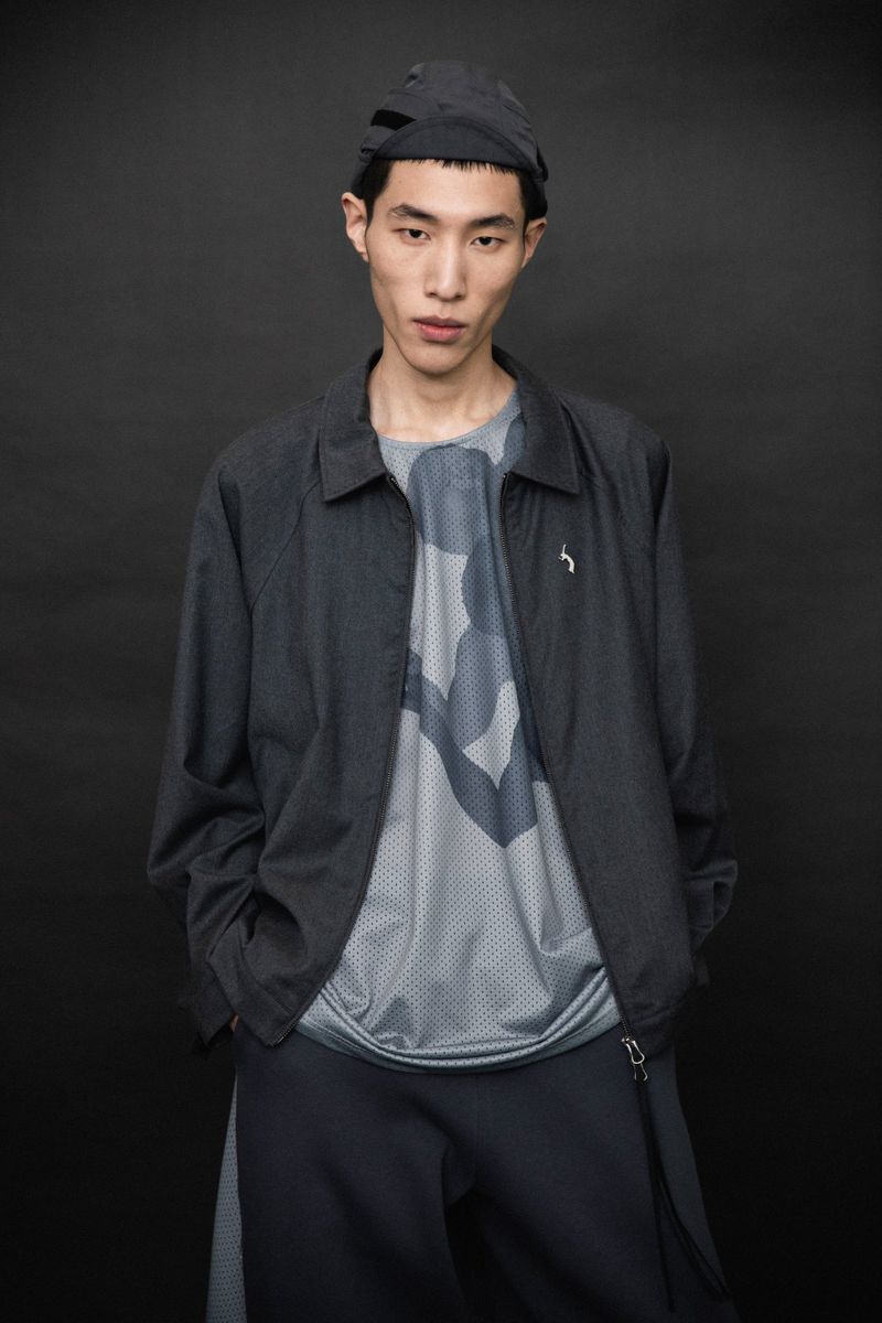 Young man in a dark athletic jacket and hat poses against a dark backdrop, casting a neutral expression in a studio setting.