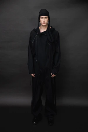 Model in a black hooded jumpsuit and matching cap standing against a dark seamless background for Saul Nash A/W 25.