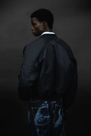 Model wearing a dark bomber jacket and patterned denim trousers seen from behind against a dark moody background