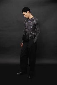 Model wearing a patterned long-sleeve top and black trousers against a dark background