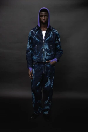 Model wearing a blue patterned jacket and matching trousers with a purple hoodie against a dark background for Saul Nash A/W 25.